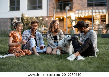 Young friends having fun playing with a dog and drinking wine, sitting on the green lawn at backyard of the country house in the evening Royalty-Free Stock Photo #2012730110