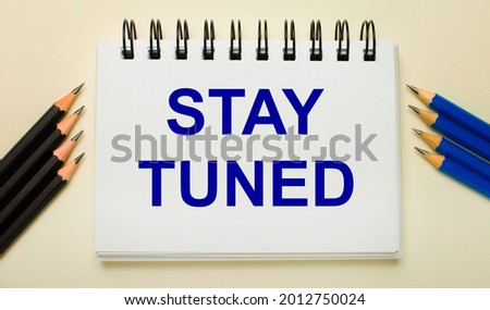 On a light background, a white notebook with the text STAY TUNED and black and blue pencils on the sides.