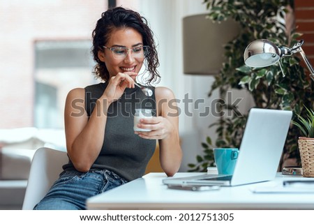 Shot of beautiful young business woman working with laptop while eating yogurt in living room at home. Royalty-Free Stock Photo #2012751305