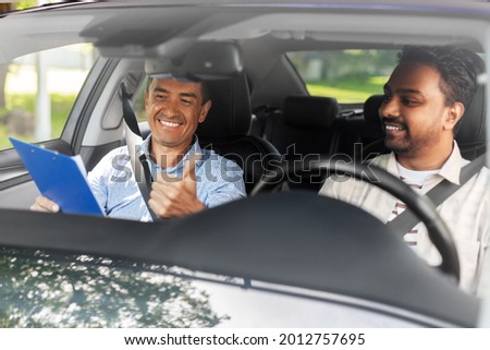 driver courses, exam and people concept - happy smiling indian man and driving school instructor with clipboard showing thumbs up in car Royalty-Free Stock Photo #2012757695