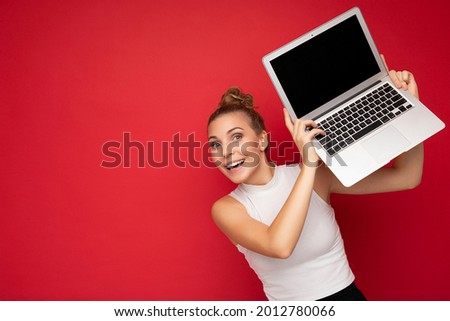 beautiful fascinating happy blond young woman with open mouth with gathered hair looking at camera holding computer laptop wearing white t-shirt isolated over red wall background