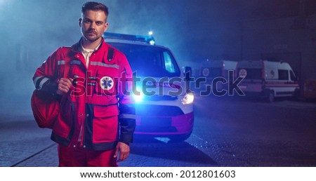 Caucasian handsome young male paramedic in red uniform getting ready to work at night shift. Ambulance car on background. Good looking man doctor outdoors with backpack. Portrait. Call 911 Royalty-Free Stock Photo #2012801603