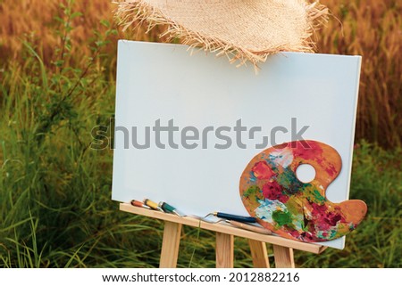 Wooden easel with blank canvas, painting equipment and hat in field. Space for text