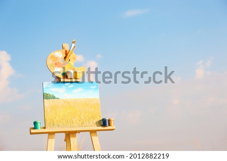 Wooden easel with beautiful picture and painting equipment against blue sky. Space for text