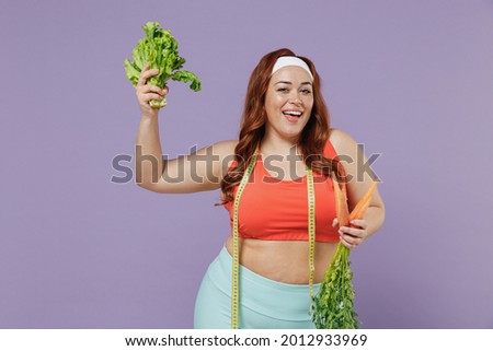 Young fun vegeterian chubby overweight plus size big fat fit woman in red top measuring tape warm up training hold carrot bunch lettuce leaves isolated on purple background gym. Workout sport concept.