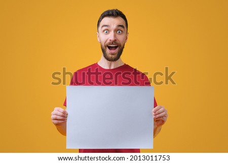 Portrait of excited bearded man holding blank white poster in hands isolated over bright colored yellow background. Mock up, copy space for yout text.
