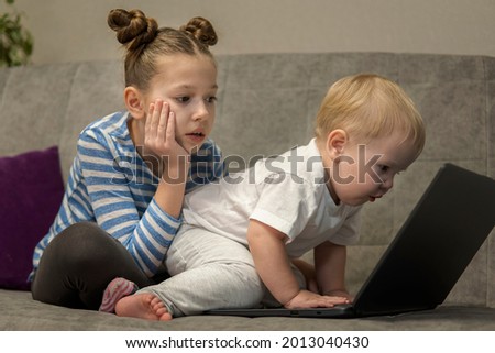 Little cute boy and girl using laptop together, looking at screen, watching cartoons or playing online, sister and brother, siblings sitting on comfortable couch at home, children and gadget concept.