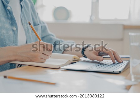Close up photo of male hands typing on computer keyboard and writing notes in agenda, freelance worker typing message and contacting client by internet from home while sitting at kitchen table