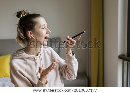 Young caucasian woman using mobile phone and wireless headphones, resting, sitting on bed at home and listening podcast or clubhouse - voice-only social media app, drop-in audio chat