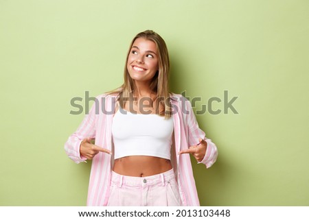 Image of attractive blond woman with flat belly, wearing stylish pink outfit, pointing fingers at stomach and looking at upper left corner with pleased smile Royalty-Free Stock Photo #2013103448