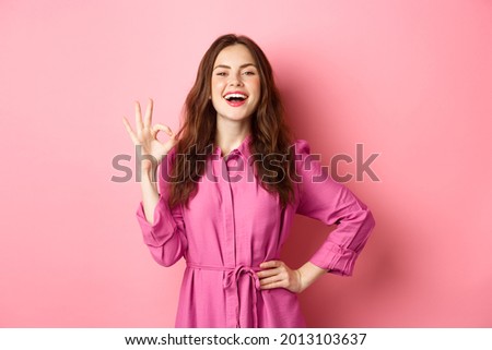 Young attractive woman saying yes, showing okay sign in approval, smiling confident and satisfied, standing over pink background