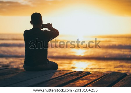 back of a sitting photographer man on a boardwalk at the beach making pictures with mobile phone during sunset