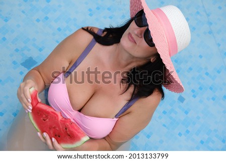 beautiful adult woman in a swimsuit, sunglasses and a summer hat holding a watermelon in the pool
