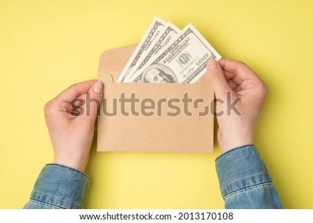 First person top view photo of female hands holding open craft paper envelope with hundred dollars banknotes on isolated yellow background with copyspace