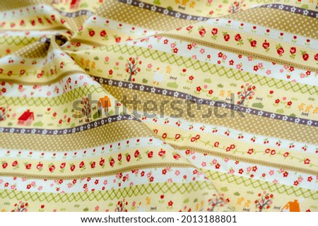 Cotton fabric, yellow shades, for children. Against the background of the fabric, stripes with a print of drawings: houses, apple trees, small flowers and ducklings.