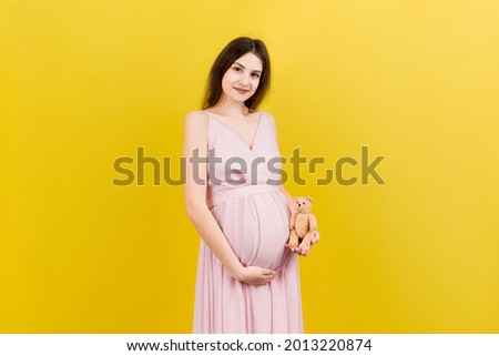Happy pregnant woman holding a teddy bear against her belly at Colored background. Young mother is expecting a baby. Copy space.
