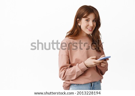 Image of smiling teenage girl with red hair, chatting on phone, messaging with smartphone and looking friendly at camera, standing over white background