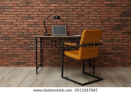 Comfortable office chair and desk with modern laptop near brick wall. Interior design