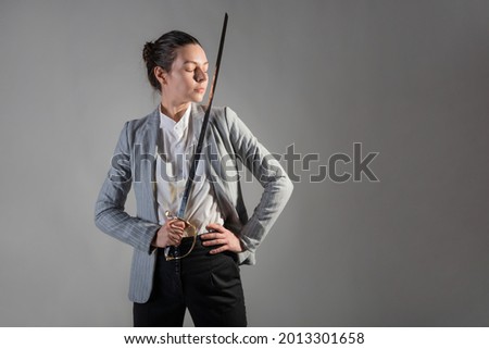 Anti-crisis management, fighting the crisis and problems at work, concept. A young businesswoman on a gray background uses a sword and fights with career challenges and solves work problems