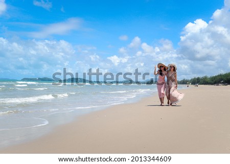 Two beautiful Asian women with big hat and sunglasses enjoy spending time with friend on tropical sand beach blue sea together, go for a walk, resting and relaxing on summer holiday vacation. 