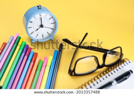 Selective focus on the alarm clock.  Colorful pencils, eyeglasses and notebook on the yellow background
