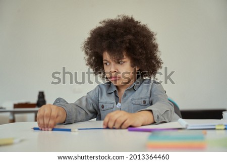 Disgruntled little schoolboy with afro hair looking sad while sitting at the table in elementary school classroom