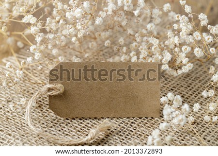 Vintage blank brown tag or card with dried white flowers on jute fabric background. Romantic love, holiday or birthday retro concept. Copy space