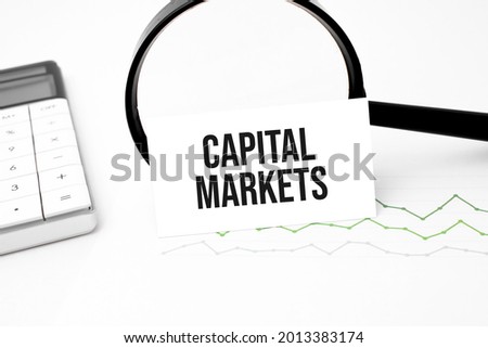 Business concept. Top view of calculator, magnifier, pen, table clock and notebook written Capital Markets on wooden background.