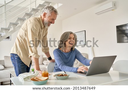 Happy middle aged 50s family couple having fun using laptop computer technology at home. Smiling senior older mature husband and wife laughing shopping online or having virtual meeting in living room.