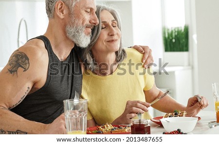 Happy affectionate mature adult husband embracing older wife enjoying having morning breakfast together at home. Mid age 50s romantic couple bonding, hugging eating waffles sitting at kitchen table.