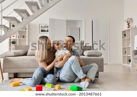 Happy young family with boy child playing enjoying time together at home in living room, parents with child sitting on floor near sofa, mom and dad holding and kissing boy, laughing and having fun. Royalty-Free Stock Photo #2013487661