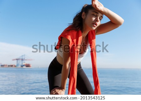Outdoor shot of tired young sportswoman having a break near the sea, wiping sweat and panting after running