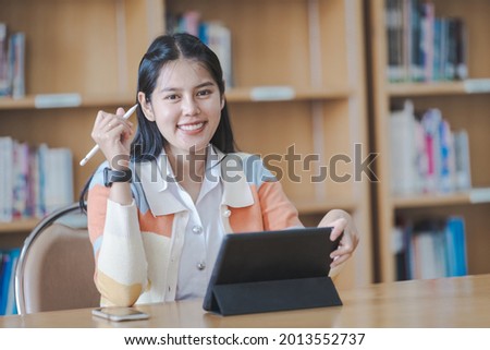 Young woman Asian college student in student uniform studying online, reading a book, using digital tablet or laptop in university library while classroom being restricted during COVID-19 pandemic