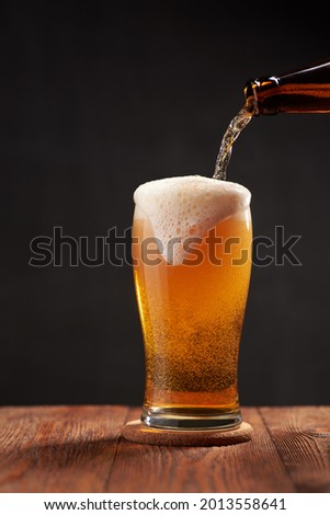 Frothy and amber brilliance craft beer, pouring in the glass from the bottle on a wooden bar.  Dark background. Shallow depth of field photo with copyspace. Royalty-Free Stock Photo #2013558641