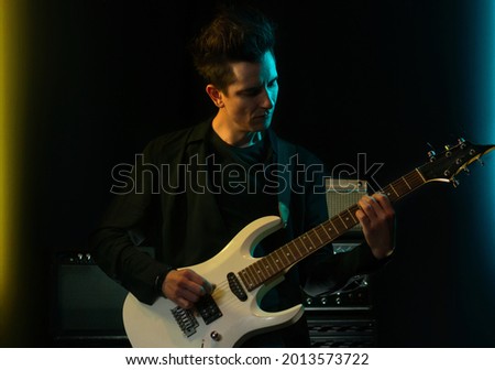 Handsome young male musician with electric guitar in recording studio. Rock musician performance in dark background with light neon effect.