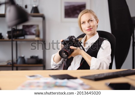 Thoughtful female photographer holding camera in hands and looking aside while sitting at office desk. Concept of people, creativity and hobby.
