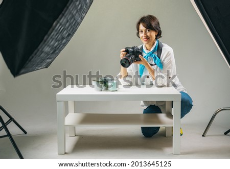 Beautiful middle aged woman in casual clothes using digital camera for making creative photos at studio. Female photographer taking pictures of cups and candles on white table.