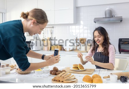 Beautiful woman pastry chef kneading bread dough on the worktop, as another female snaps a snapshot of her doing so.