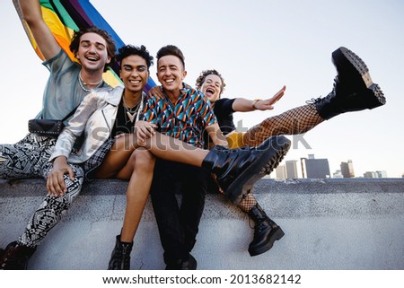 Four LGBTQ people celebrating pride while sitting together. Four friends smiling cheerfully while raising the rainbow pride flag. Group of young queer individuals celebrating together outdoors. Royalty-Free Stock Photo #2013682142