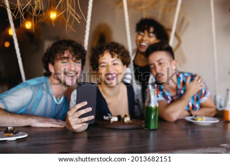 Group of friends taking a selfie together in a restaurant. Four young queer people smiling cheerfully while capturing the moment. Friends sitting and bonding together during lunch. Royalty-Free Stock Photo #2013682151