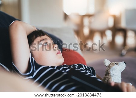 Side view portrait young boy sitting on sofa watching TV, Positive Kid lying on couch with dog toy in living room. Child relaxing at home on weekend in Spring or Summer 