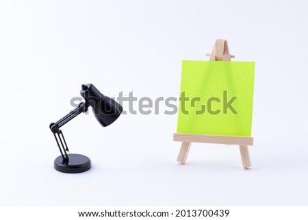 Wooden Easel Miniature with Blank Colored Square Canvas or Memo Paper - Mockup. Mini Wooden Stand with Clean Artboard and Small Black Table Lamp on White Background, Copy Space
