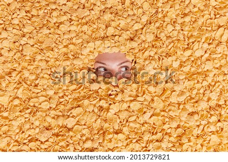 Top view of young woman fully covered in cereals focused away buried in cornflakes makes creative shot with food going to have breakfast. Healthy balanced nutrition concept. Horizontal shot.