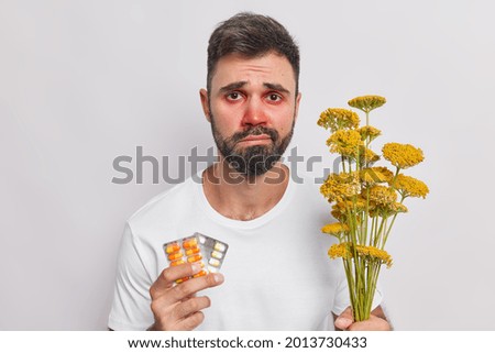 Sad man with beard red swollen eyes suffers from allergic rhinitis to flower pollen holds pills to cure disease feels unhealthy has discontent expression wears casual t shirt isolated on white wall Royalty-Free Stock Photo #2013730433