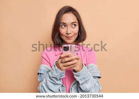 Dreamy young pretty Asian woman with dark hair holds mobile phone has thoughtful expression thinks about received message wears pink t shirt denim jacket looks away isolated over beige background. Royalty-Free Stock Photo #2013730544