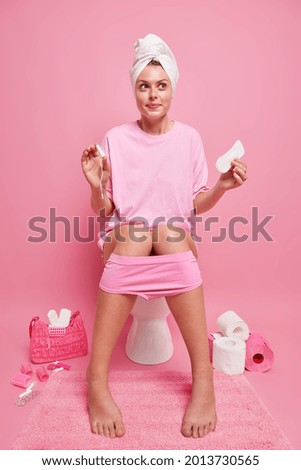 Full length shot of pretty thoughtful woman wears casual t shirt towel over head comfortable panties holds menstruation cotton tampon and sanitary napkin poses on toilet bowl keeps feet on carpet