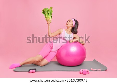 Fitness woman leans on swiss ball holds fresh green vegetable keeps to heathy diet has regular training at home to keep fit isolated over pink background. People proper nutrition and sport concept