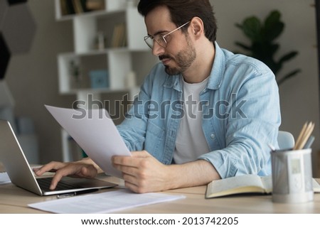 35s businessman sit at desk make online research, paperwork, work online on laptop, holds bills looks at computer screen pay bills via e-bank app typing at modern device, prepare audit report concept Royalty-Free Stock Photo #2013742205