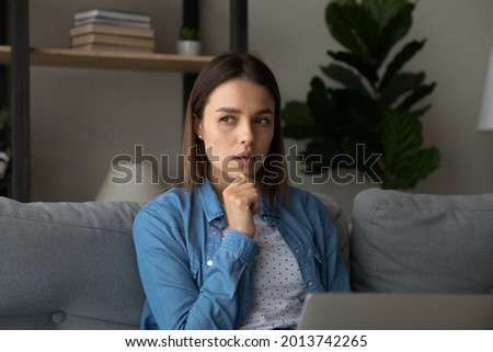 Pensive young woman sit on couch looking deep in thoughts touch chin with hand while thinks over freelance task working remotely from home on laptop. Thoughtful female studying use modern tech concept Royalty-Free Stock Photo #2013742265
