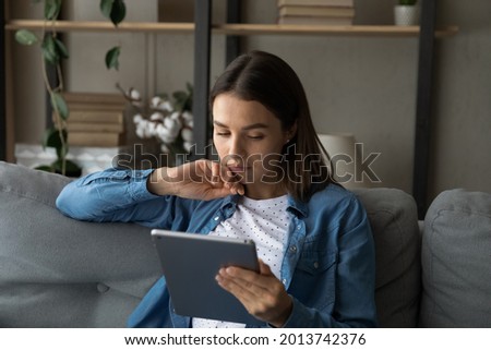 Young serious woman sit on sofa spend free time use modern wireless tech, read media news on tablet looking serious or pensive, lean new mobile app, choose goods ponder over telecommute task concept Royalty-Free Stock Photo #2013742376